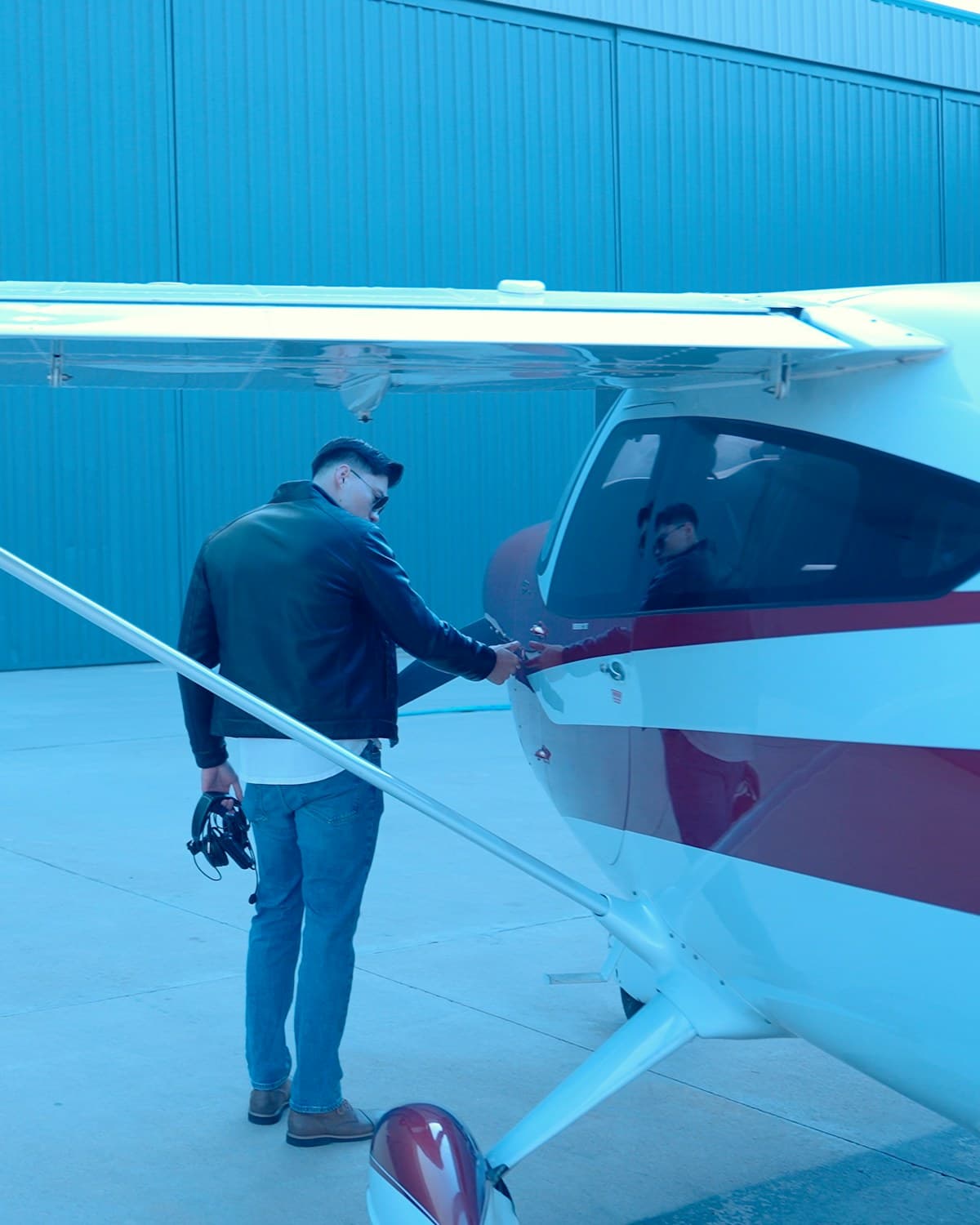 Hombre junto a una avioneta blanca y bordó, con auriculares en la mano, tocando la puerta de la cabina frente a un hangar azul.