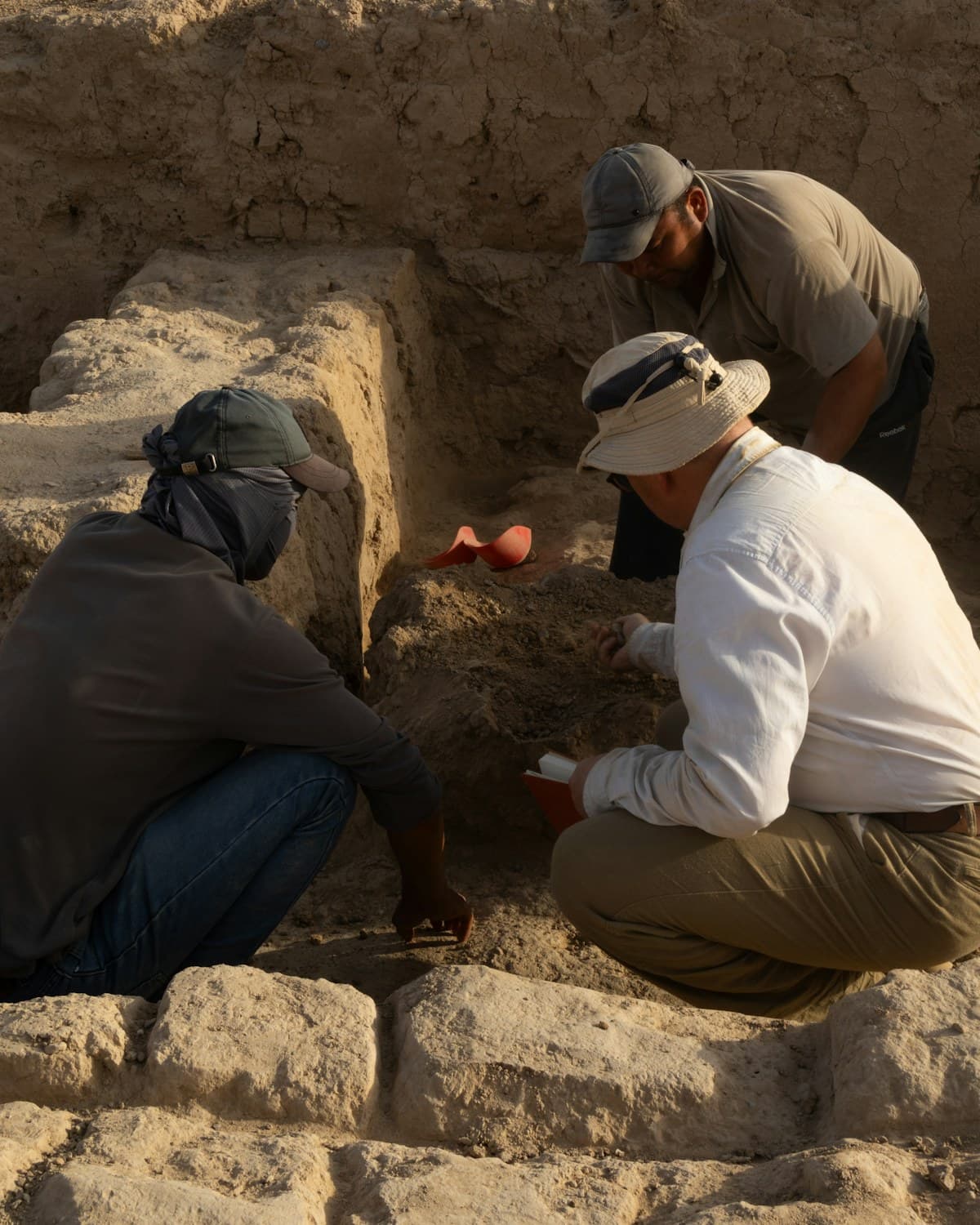 Tres personas trabajan agachadas en una excavación de tierra junto a muros de piedra, con pequeñas herramientas de mano.