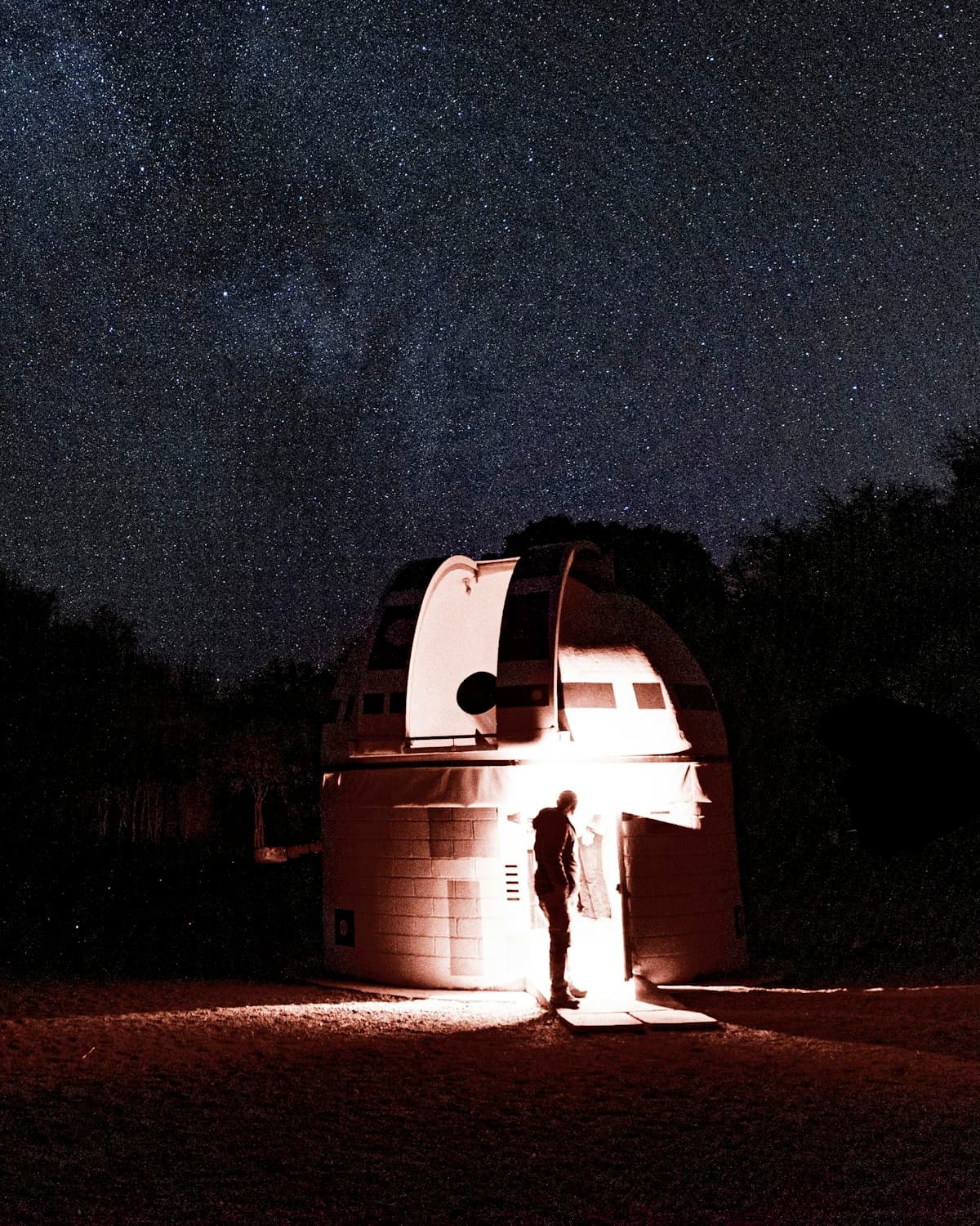 Una persona junto a la entrada iluminada de un observatorio con cúpula abierta, bajo un cielo estrellado.