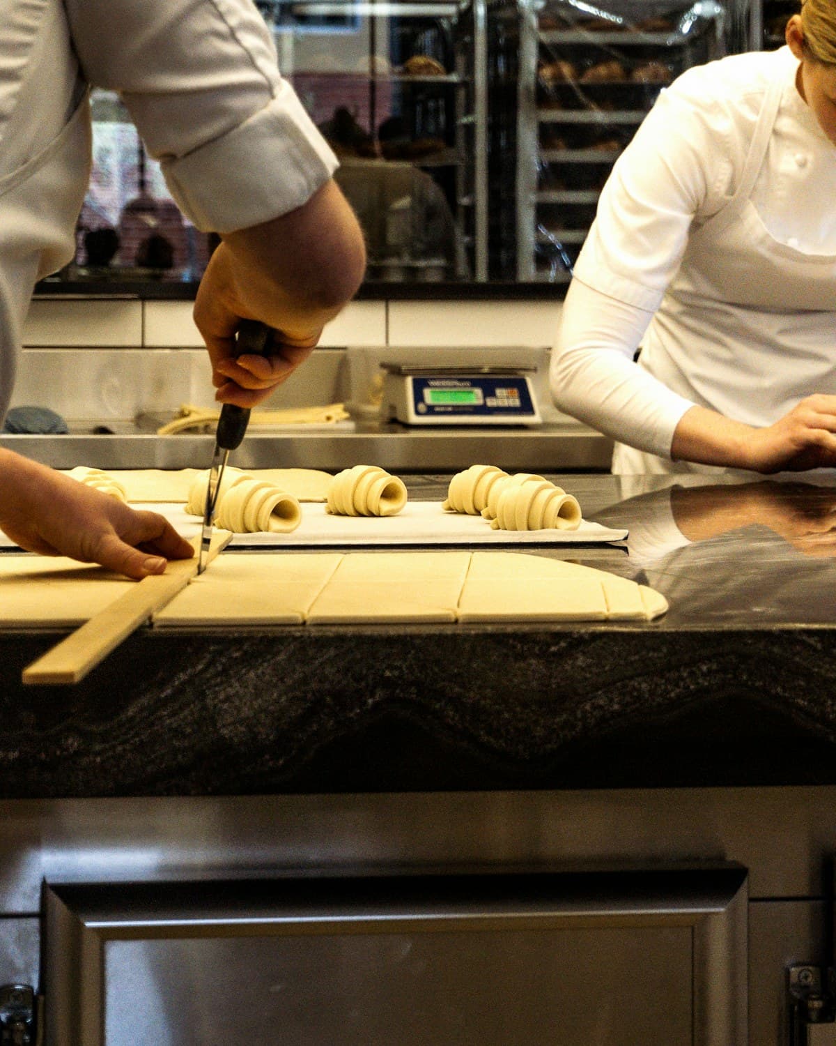 Dos personas con chaquetas blancas preparan masa enrollada sobre una mesada de cocina, junto a bandejas y utensilios.