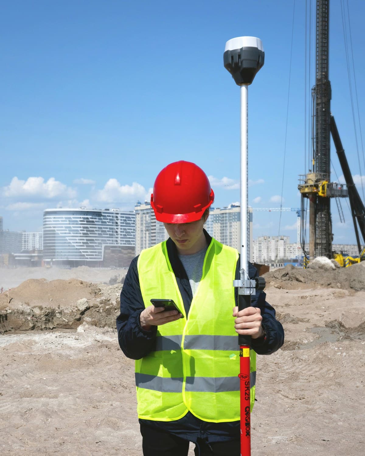 Persona con casco rojo y chaleco reflectante sostiene un receptor topográfico y un teléfono en una obra, con maquinaria y edificios al fondo.