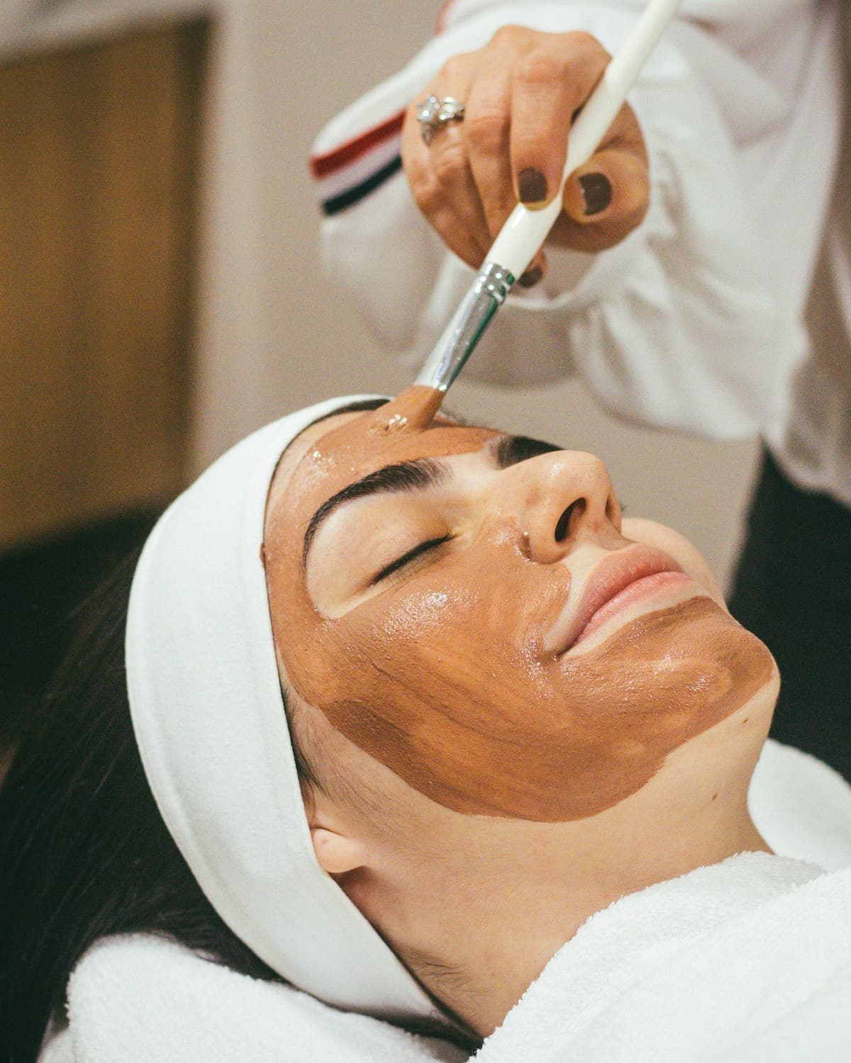 Profesional aplica una mascarilla facial con pincel a una persona recostada en una camilla.