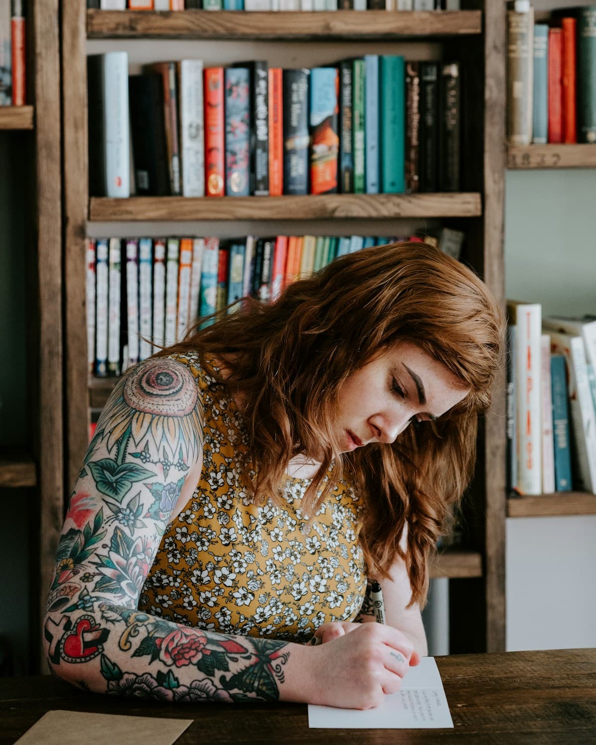 Persona escribiendo a mano en una hoja sobre una mesa, con estantes llenos de libros al fondo.
