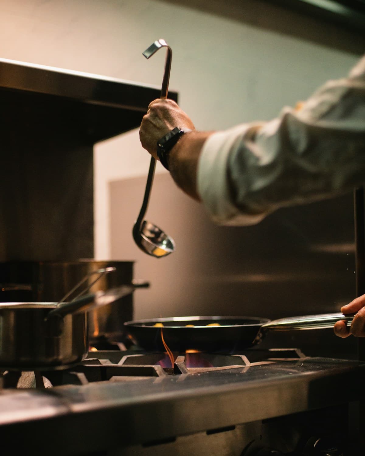 Persona con chaqueta de cocina sostiene un cucharón sobre una sartén con llama en una cocina profesional.