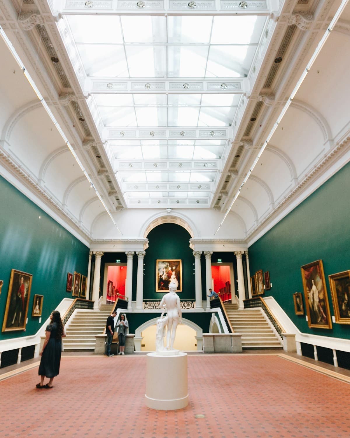 Sala de museo con una escultura blanca en el centro, escaleras laterales y pinturas colgadas en paredes verdes.