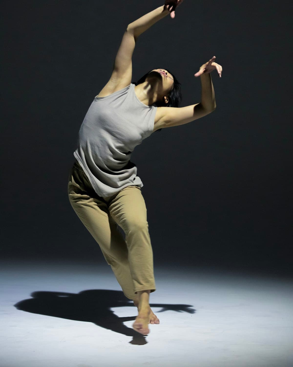 Bailarina descalza con remera gris y pantalón beige realizando un movimiento en un estudio oscuro, sobre un piso iluminado.
