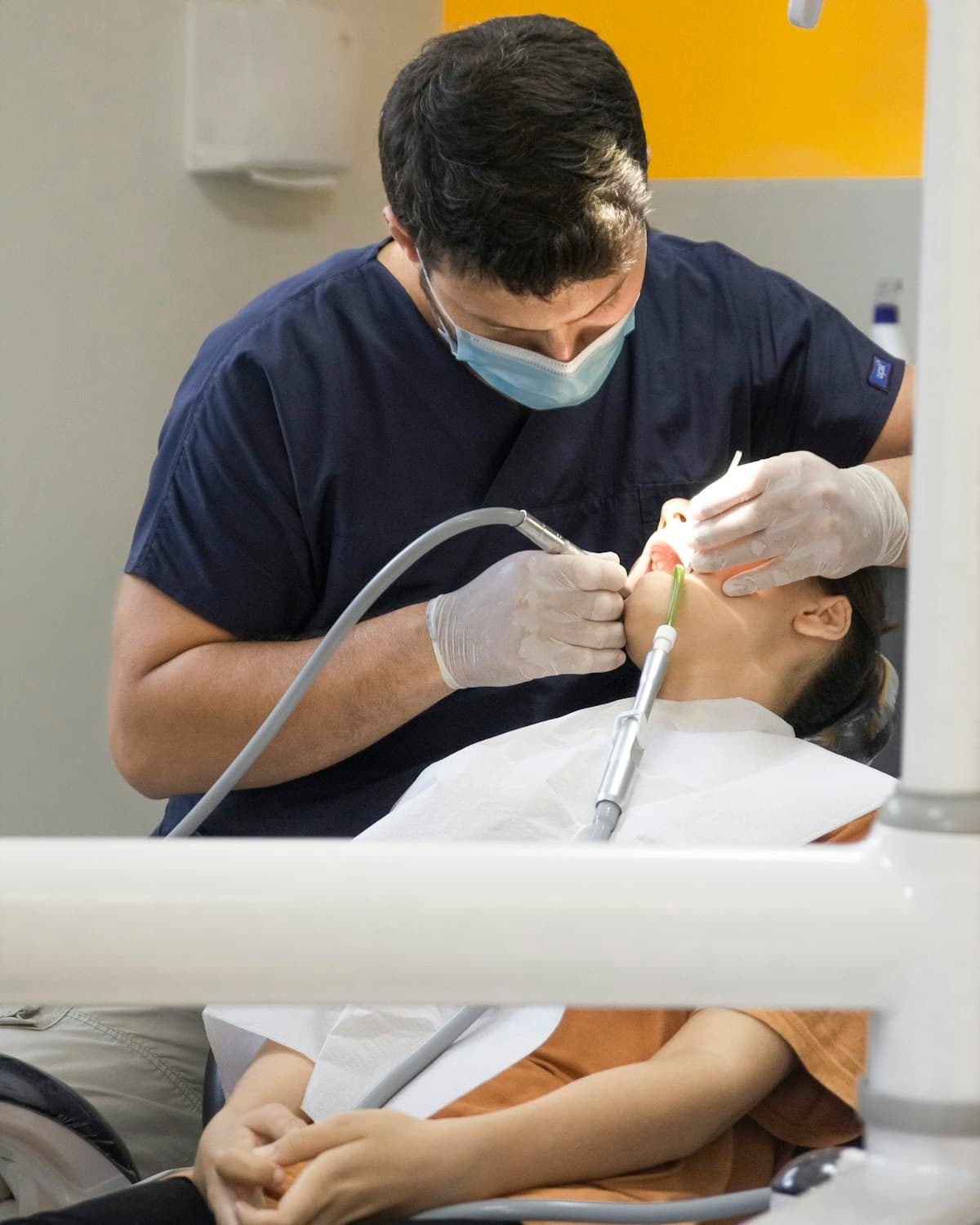 Profesional de odontología con barbijo y guantes realiza un procedimiento en la boca de una paciente reclinada en un sillón dental.