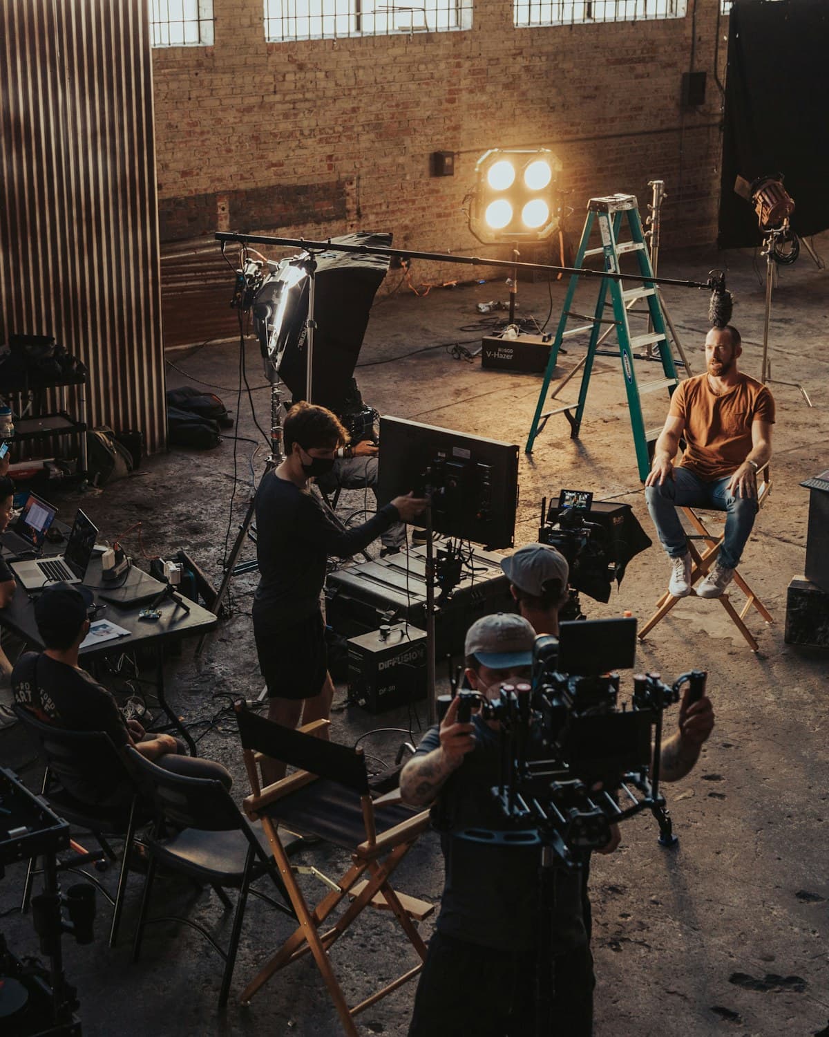 Equipo de filmación en un estudio industrial, con cámara, monitores, luces, escalera y una persona sentada frente al set.
