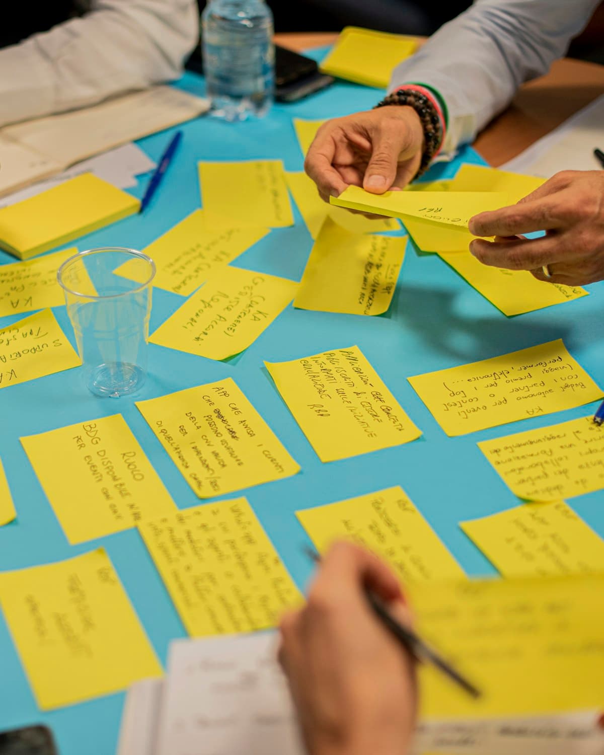 Manos de varias personas organizando notas adhesivas amarillas sobre una mesa azul, con anotaciones y vasos de plástico.