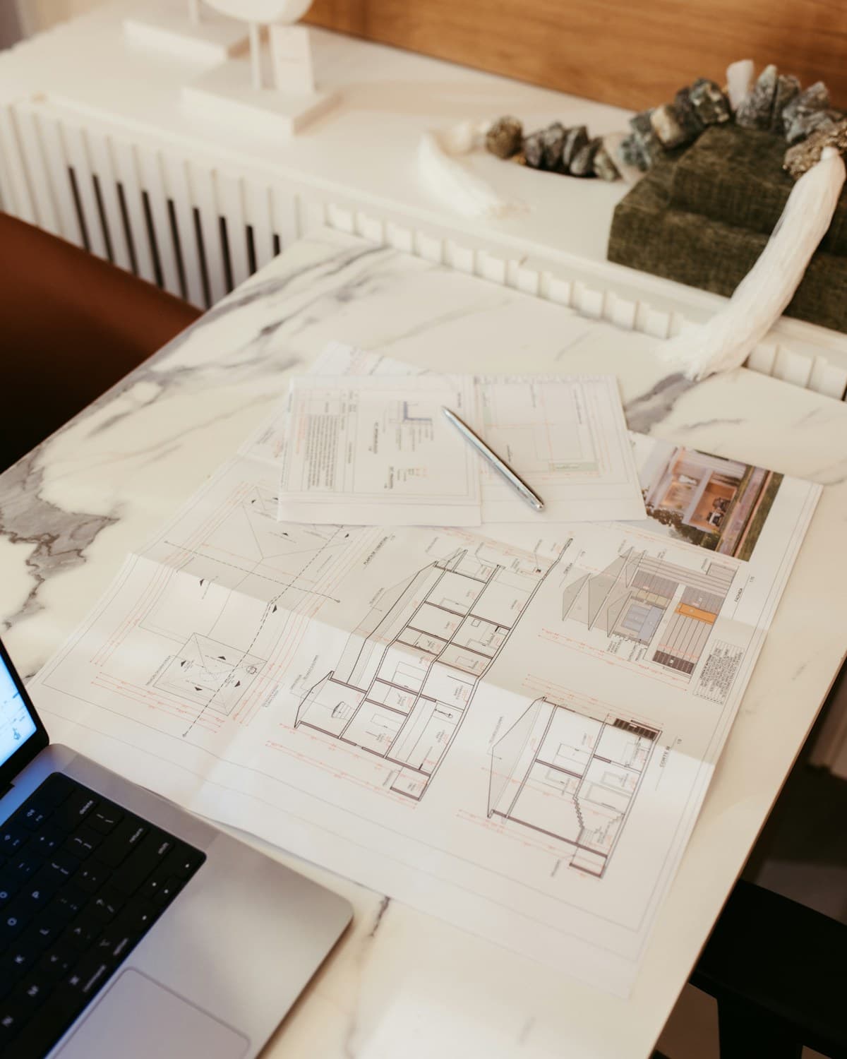 Planos impresos, una libreta con lápiz, una maqueta y una computadora portátil sobre una mesa.
