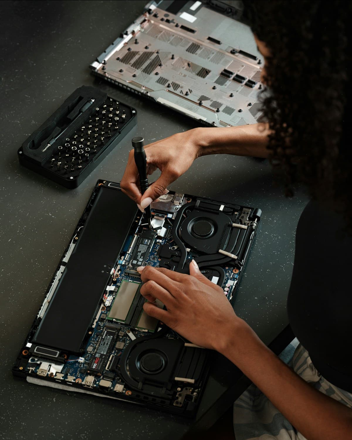 Vista cenital de una persona usando un destornillador sobre los componentes internos de una computadora portátil abierta, junto a una bandeja con tornillos.