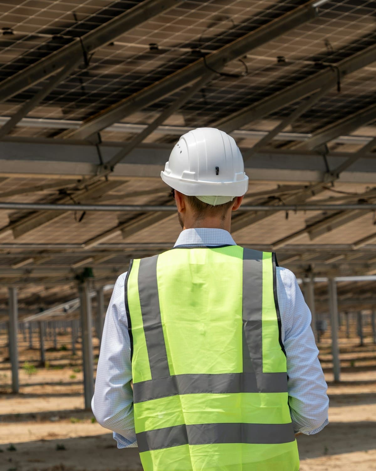 Persona con casco blanco y chaleco reflectante de espaldas bajo una estructura de paneles solares.