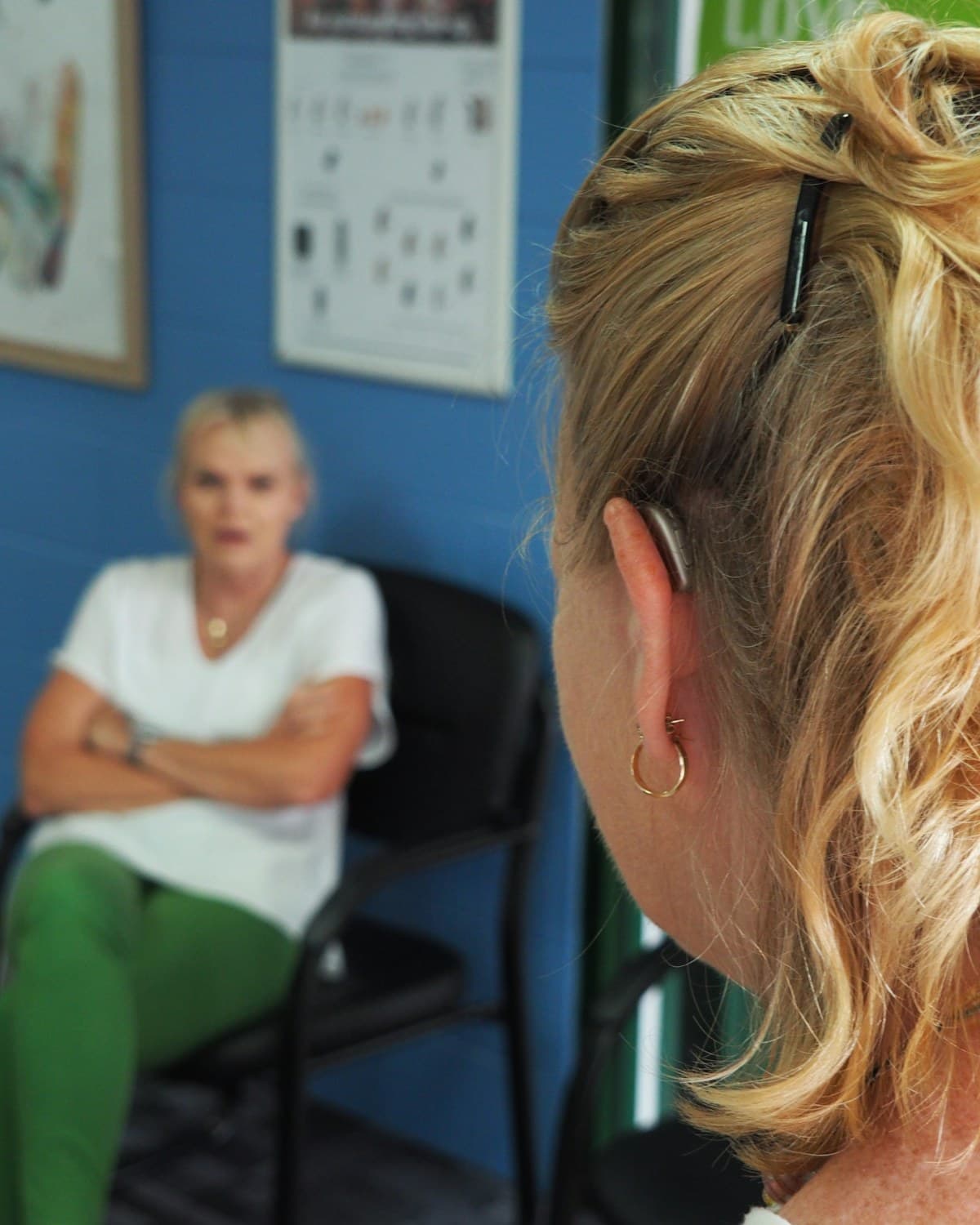Vista desde atrás de una mujer con un audífono detrás de la oreja frente a otra mujer sentada en un consultorio.