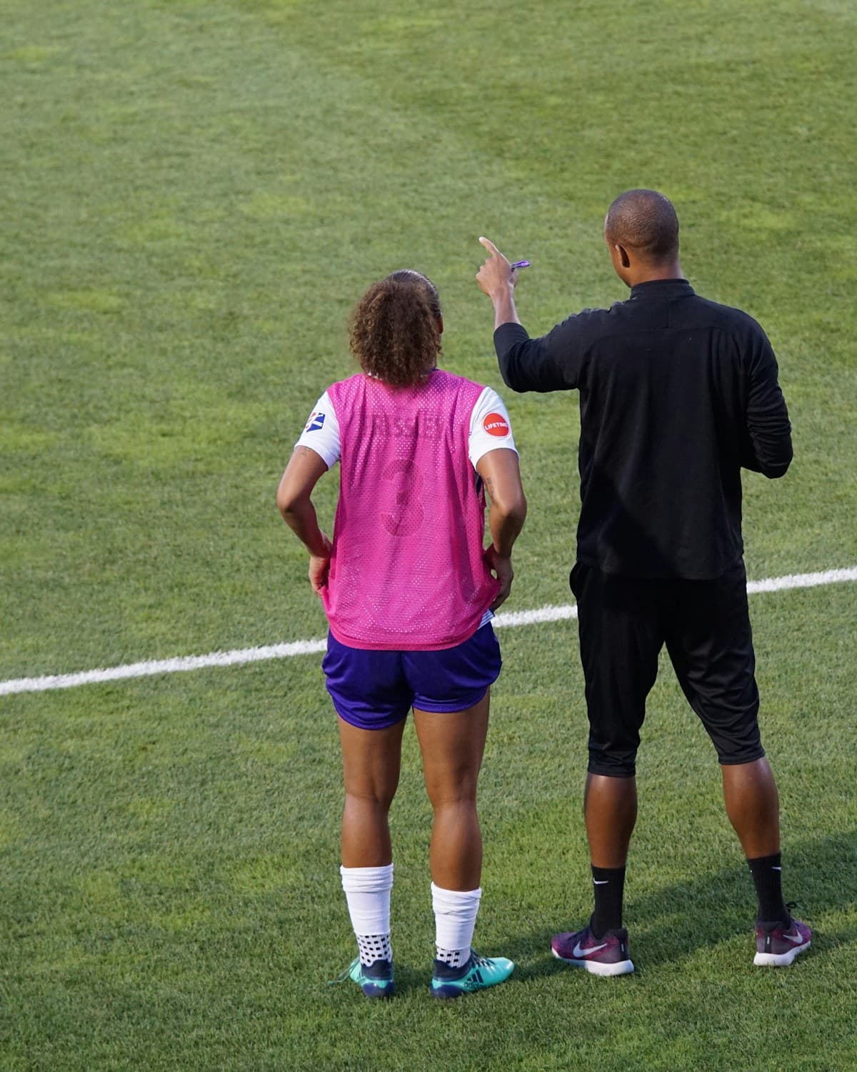 Entrenador de pie junto a una jugadora de fútbol en una cancha de césped, mientras hace un gesto con la mano.
