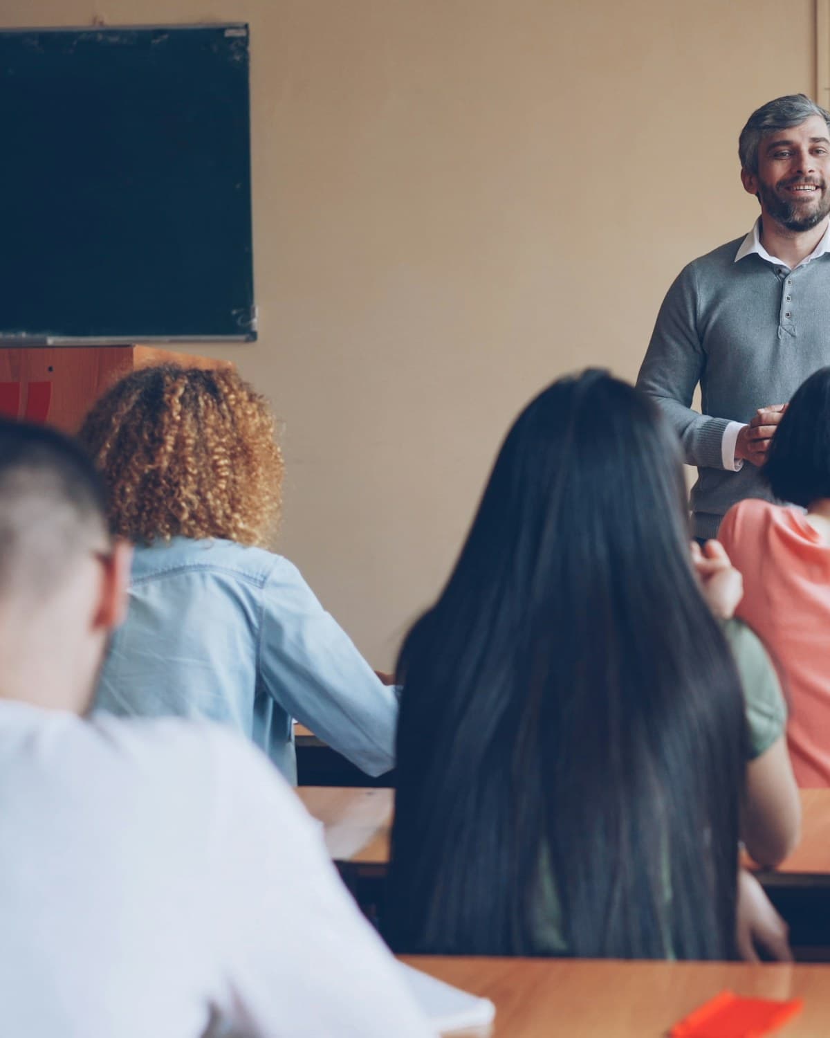 Docente de pie frente a un grupo de estudiantes sentados en un aula, junto a un pizarrón.