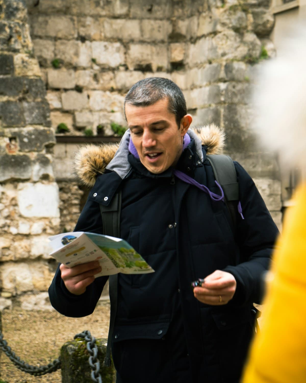 Hombre con campera y mochila sostiene un mapa junto a un muro de piedra antiguo.