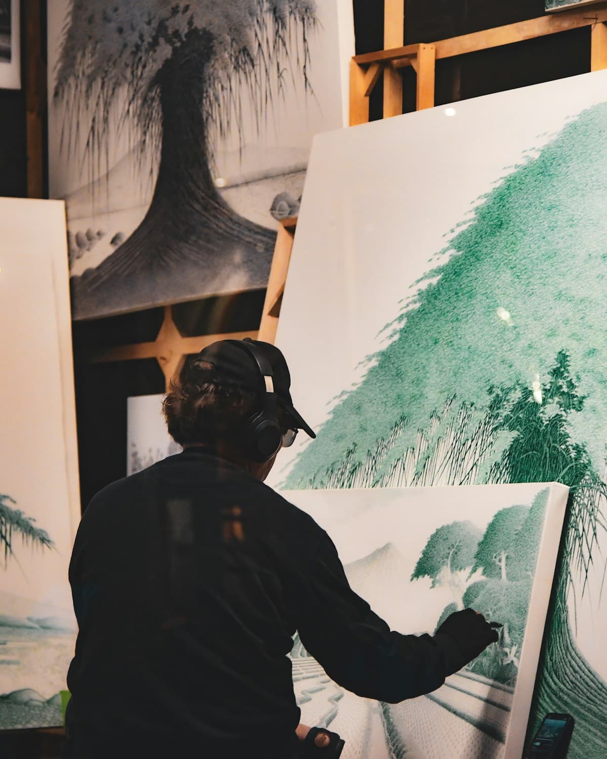 Persona de espaldas trabajando sobre una pintura en un taller, rodeada de lienzos con dibujos de árboles.