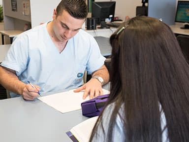 Estudiante y docente con uniforme sanitario revisan apuntes en una mesa de trabajo.