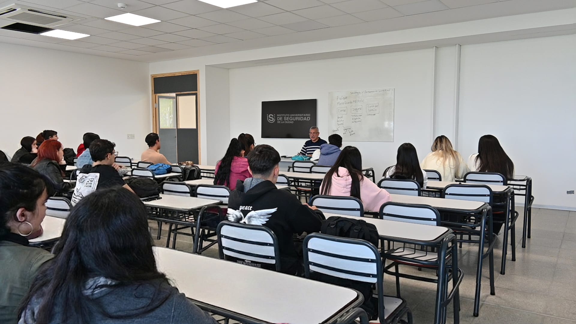 Aula con estudiantes sentados frente a una docente durante una clase.