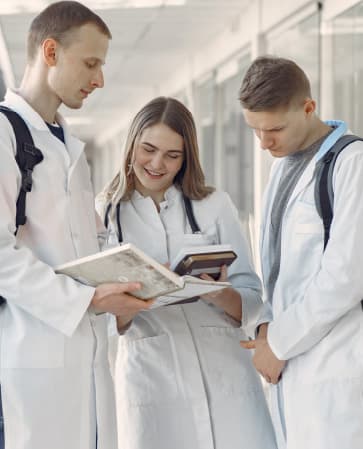 Tres personas con guardapolvos consultan material de estudio en un pasillo