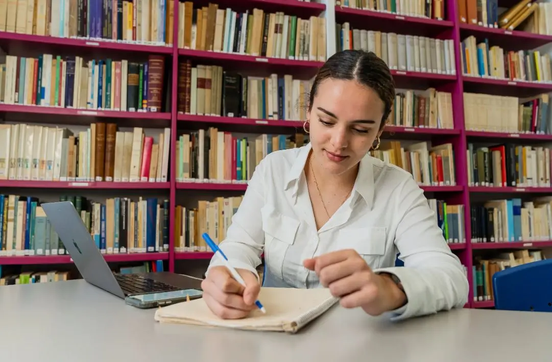 Estudiante escribiendo en un cuaderno frente a una computadora portátil y estanterías de libros