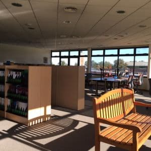 Sala de biblioteca con estanterías, mesas de estudio y un banco junto a ventanales