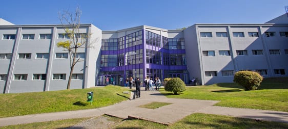 Edificio del campus con acceso central acristalado y estudiantes en el exterior.