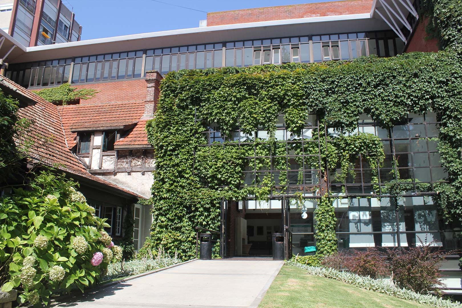Edificio universitario de ladrillo y grandes ventanales, con ingreso rodeado de vegetación.