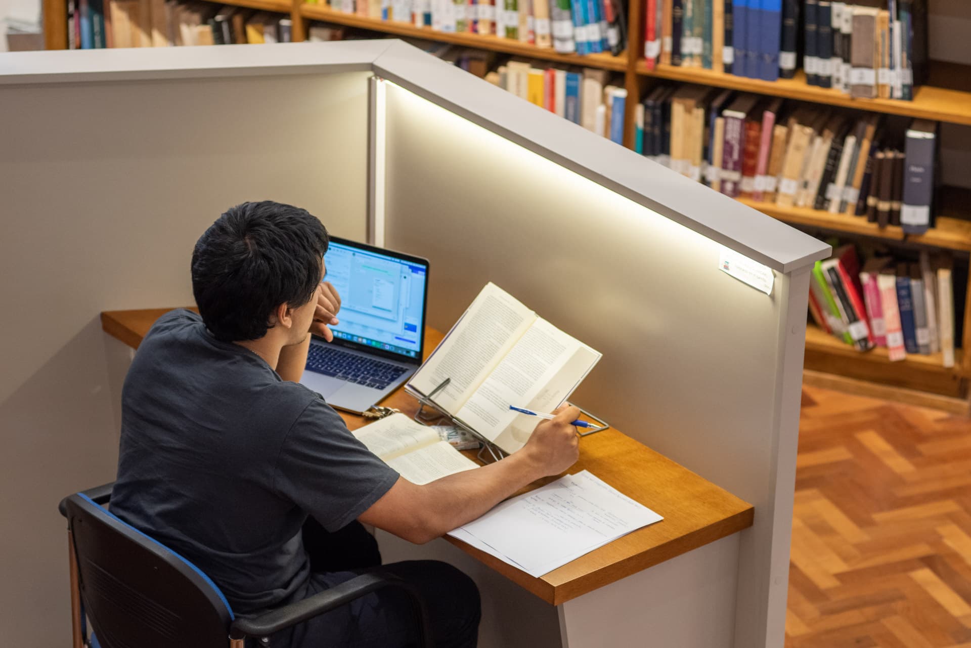 Estudiante trabajando con una computadora portátil y libros en un puesto individual de biblioteca.