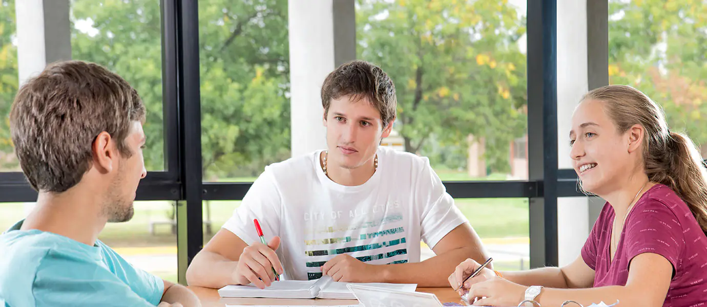 Tres estudiantes conversan y revisan materiales de estudio junto a ventanales