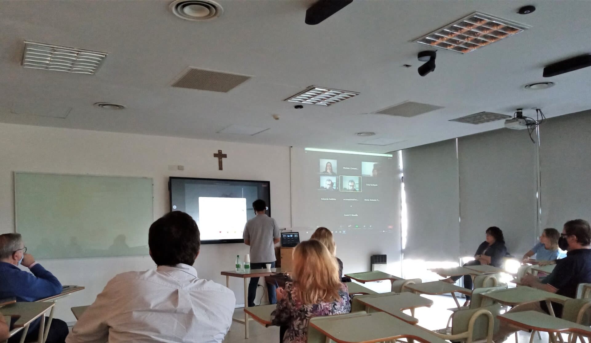 Clase en aula híbrida con asistentes y una presentación proyectada