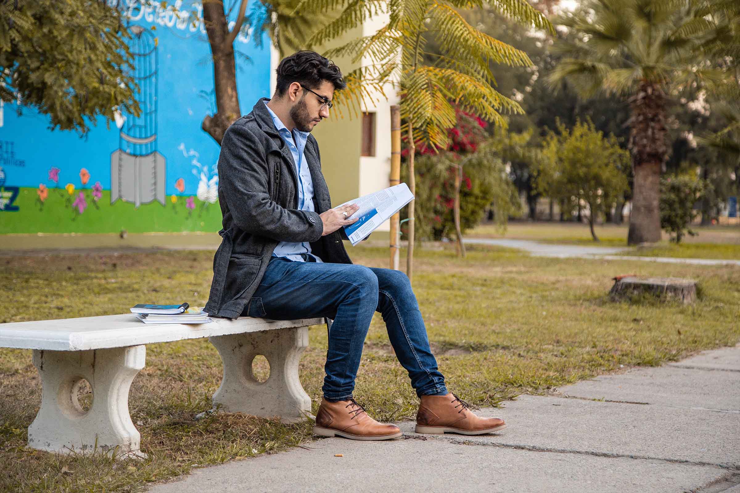 Estudiante sentado en un banco al aire libre mientras lee apuntes, junto a un sendero y edificios del campus.