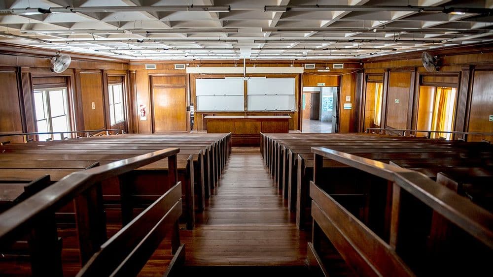 Aula escalonada vacía con bancos de madera, pizarrón y pantalla de proyección.