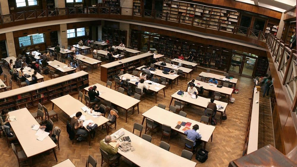 Sala de lectura de biblioteca con mesas ocupadas y estanterías de libros en varios niveles.