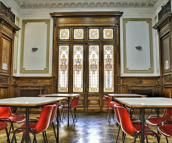 Sala de lectura con mesas, sillas rojas y vitrales.