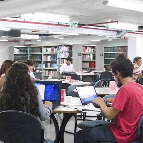 Estudiantes trabajando con computadoras en mesas de una biblioteca.