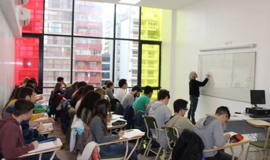 Clase con estudiantes sentados y docente frente a un pizarrón