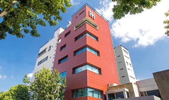 Edificio de varios pisos con fachada roja y blanca, visto desde la calle entre árboles.