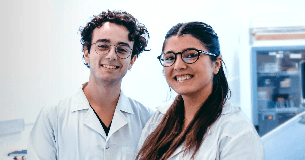 Dos personas con guardapolvos blancos en un espacio de laboratorio con equipamiento al fondo.