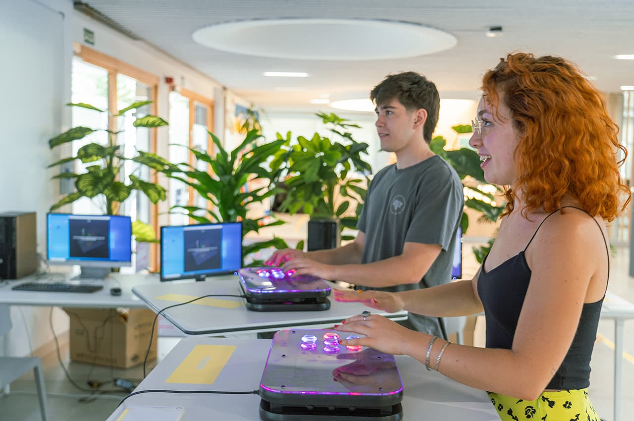 Dos estudiantes interactúan con controles luminosos junto a computadoras en un espacio interior.