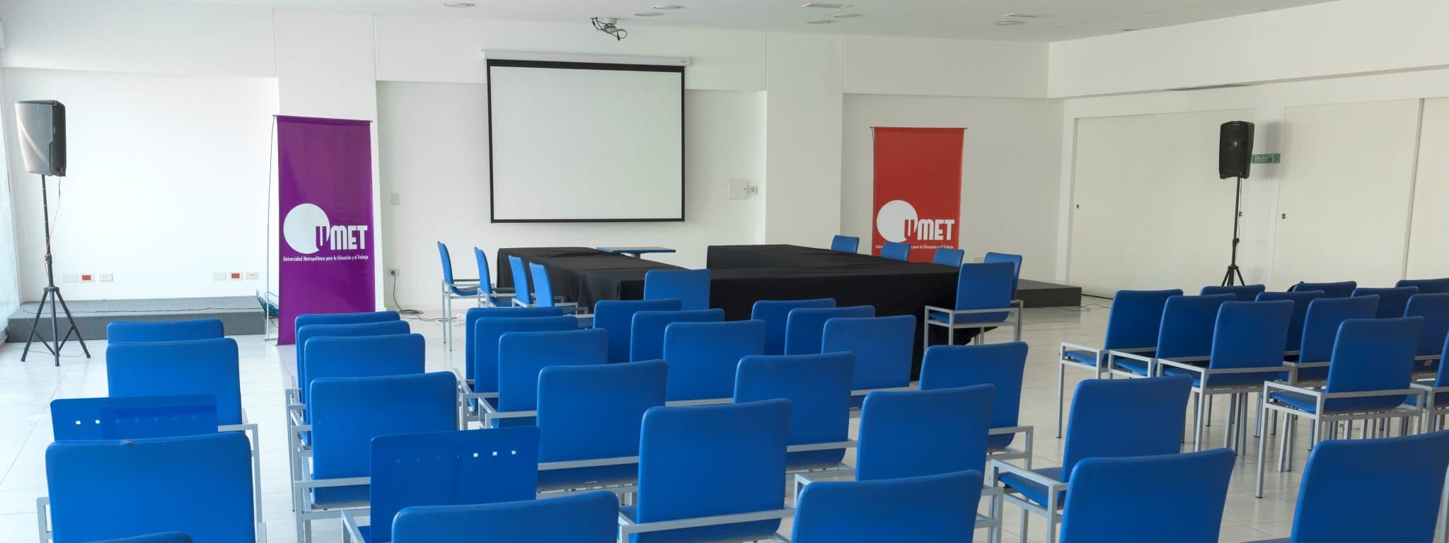 Aula o auditorio con filas de sillas azules, pantalla de proyección y mesa al frente