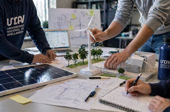 Estudiantes trabajan con una maqueta, paneles solares y planos sobre una mesa de estudio.