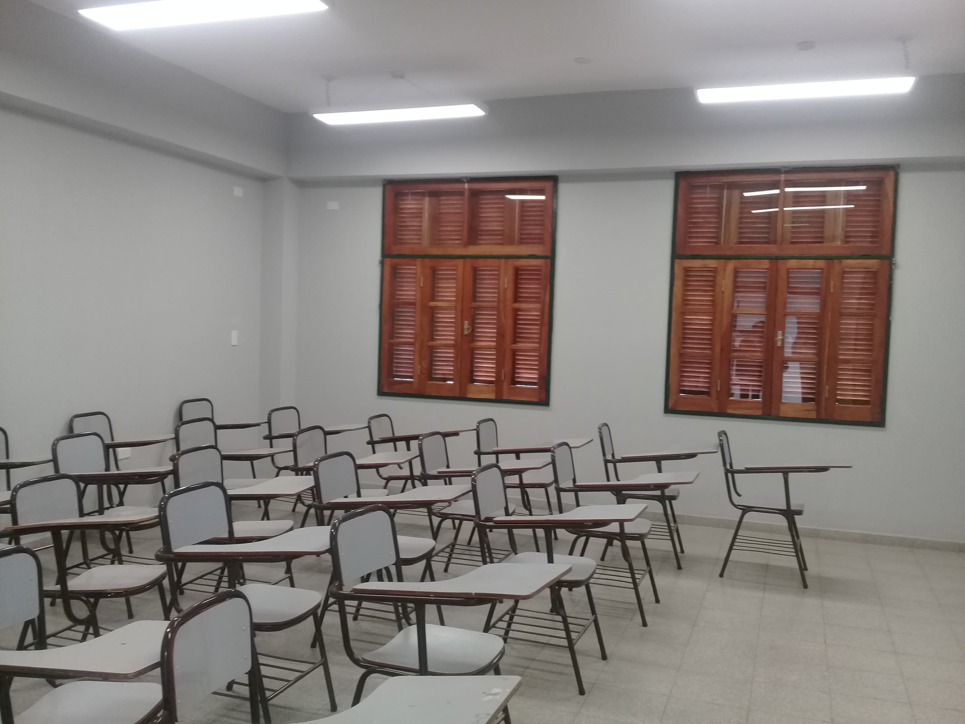 Aula con filas de pupitres, ventanas de madera y luminarias en el techo.