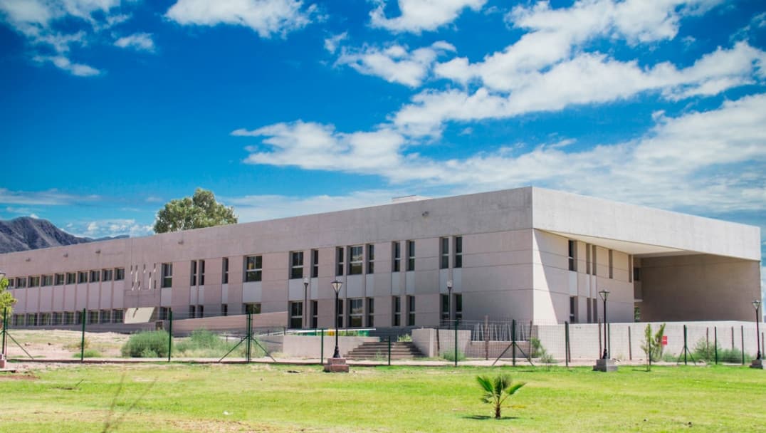 Edificio universitario de dos plantas con césped y montañas al fondo