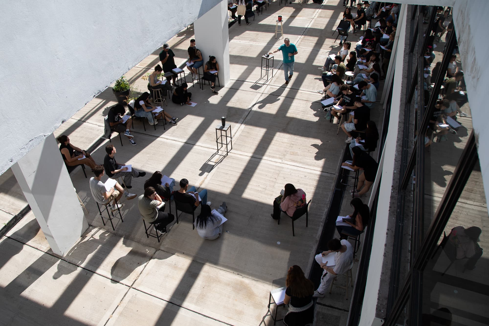 Vista elevada de una clase con estudiantes sentados en un patio interior cubierto.