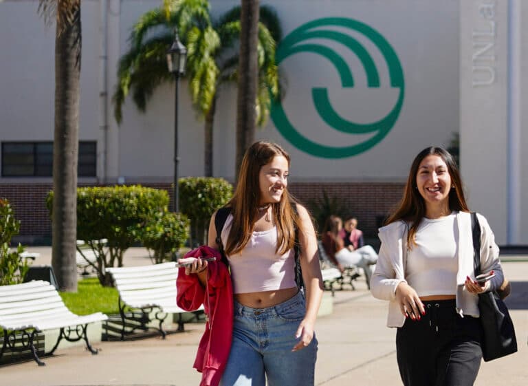 Dos personas caminan por un sendero del campus frente a un edificio con el emblema de la UNLaM.