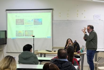 Clase en aula con docente frente a una proyección y estudiantes sentados.