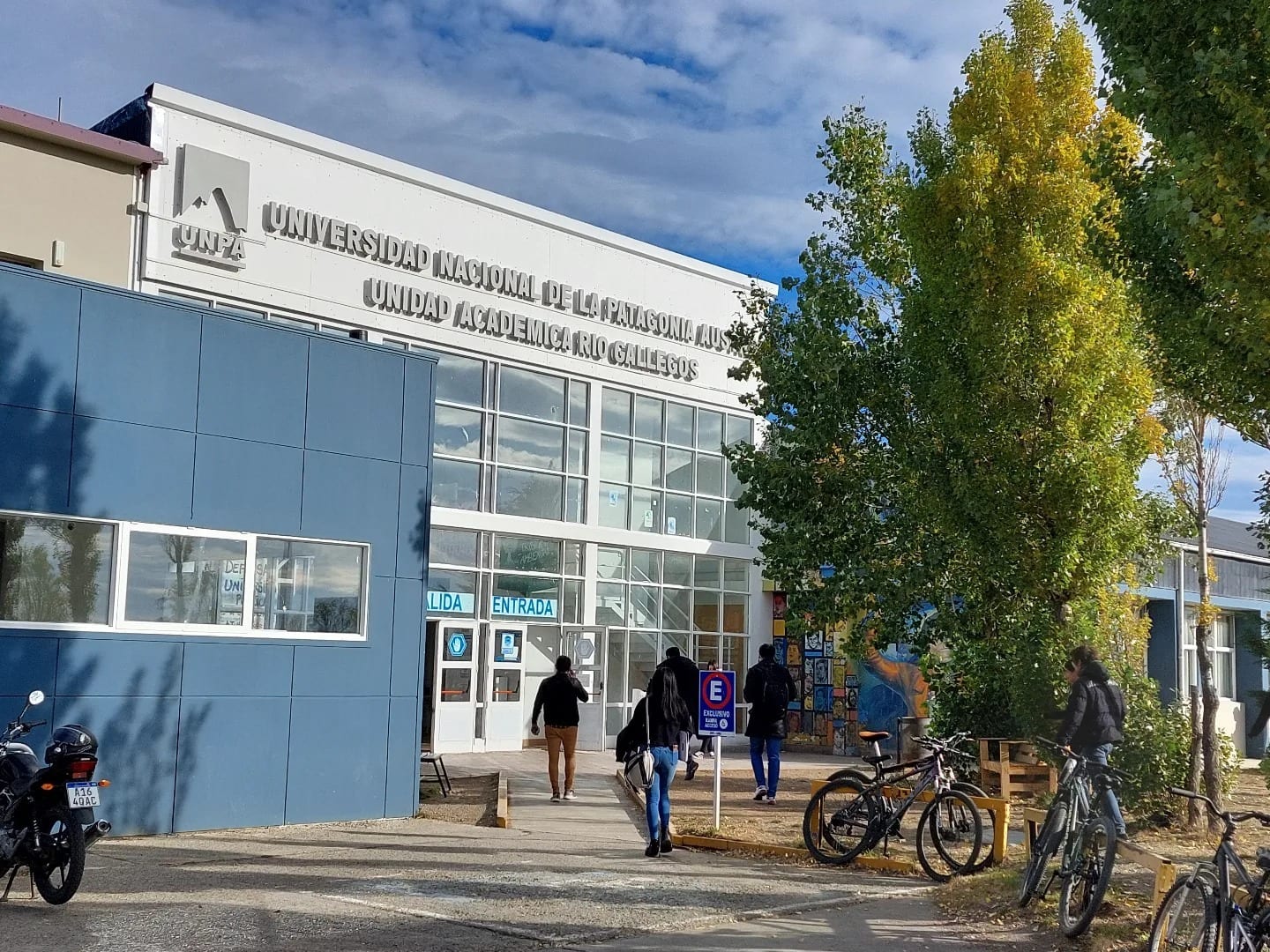 Fachada de un edificio universitario con ingreso, personas caminando y bicicletas estacionadas.