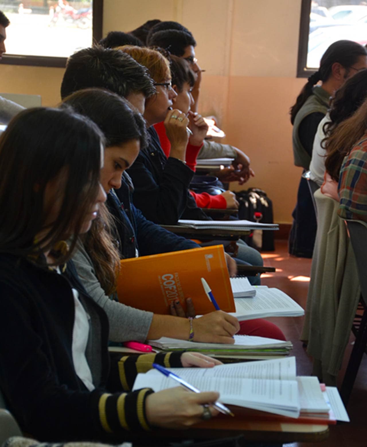 Estudiantes sentados en un aula, tomando apuntes y leyendo material de estudio.