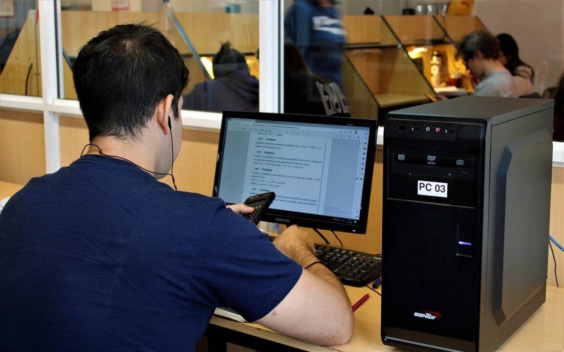 Estudiante trabaja frente a una computadora en un espacio de biblioteca con estanterías al fondo.